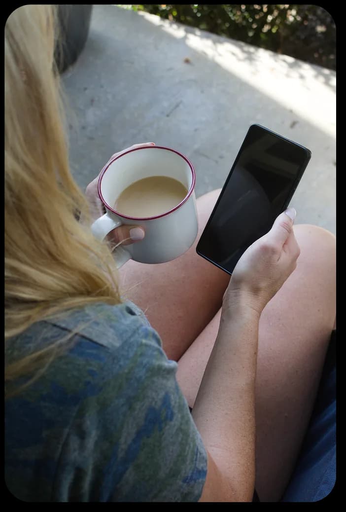 A woman on a porch holding a mug of coffee in one hand and her phone in the other, waiting to hear that her hunter made it out.