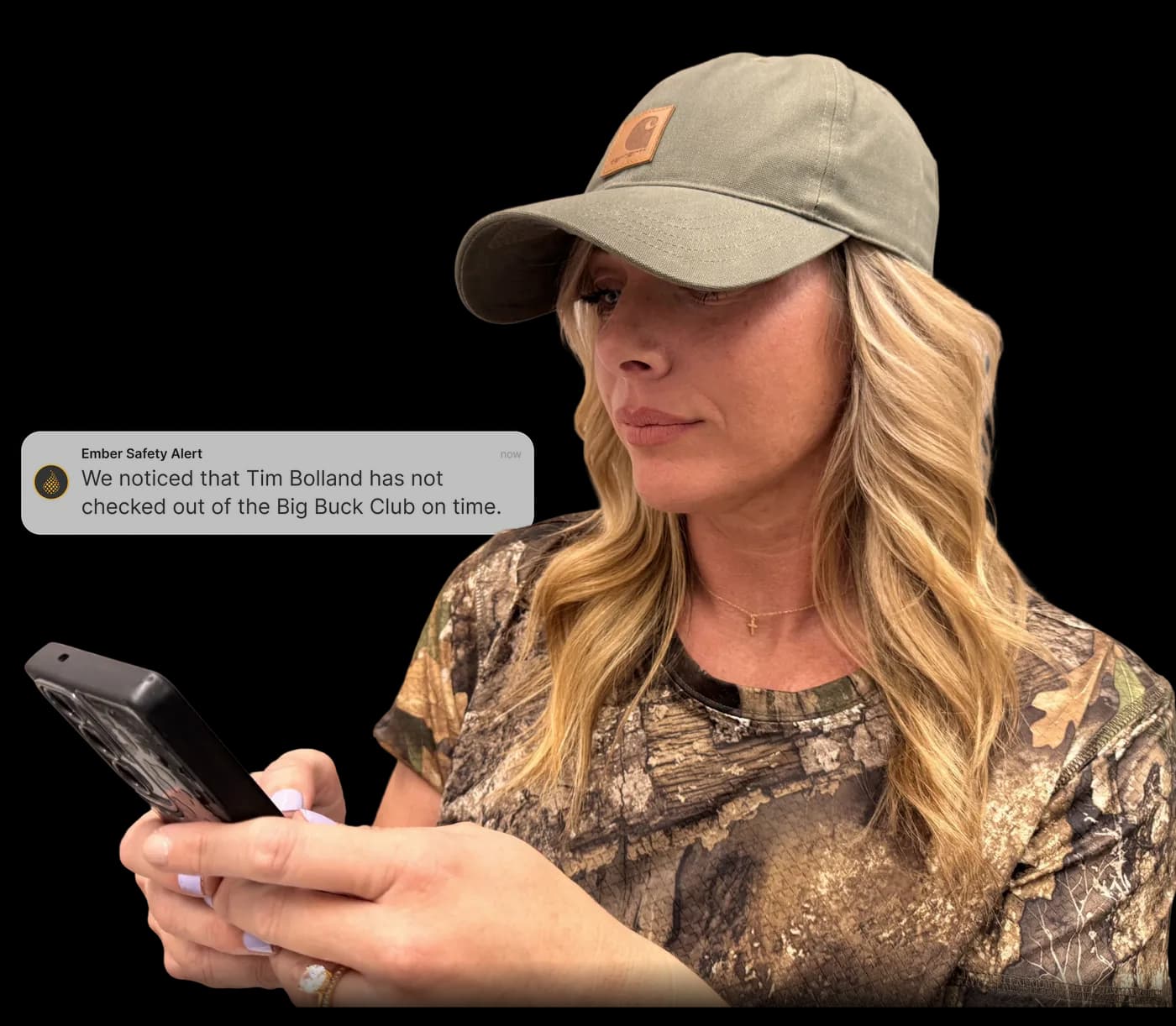 A hunter in a camo shirt and ball cap looking at her phone, which shows an Ember Safety Alert that Tim Bolland has not checked out of the Big Buck stand